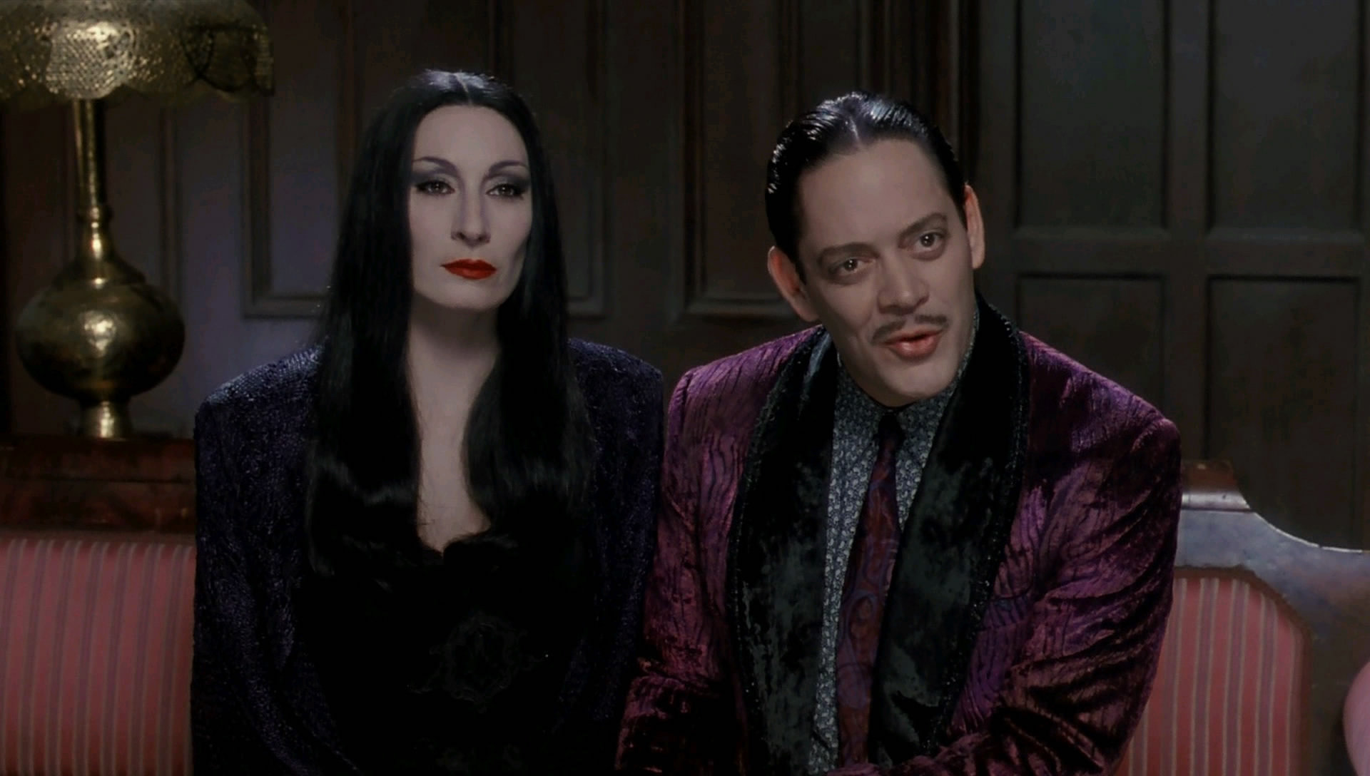 Morticia and Gomez Addams, sitting on a sofa. Gomez appearing to be in mid sentence.