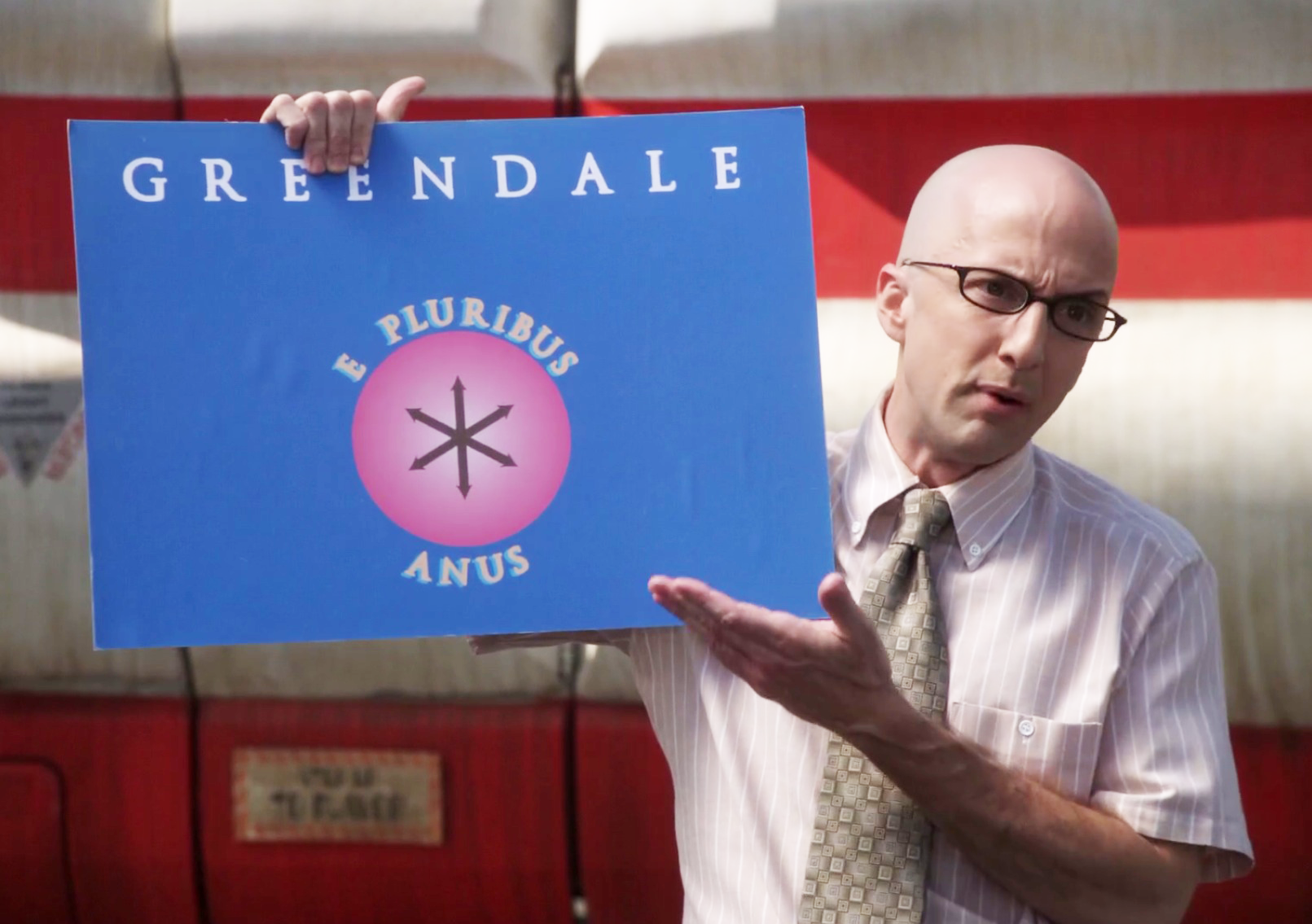 Dean Craig Pelton from Community gesturing disgustedly at the Greendale Community College flag, which has a pink asterisk logo and the motto "e pluribus anus".