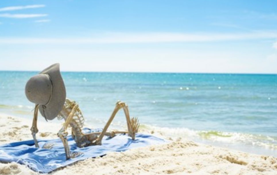 Stock image of a skeleton wearing a sunhat sitting on the beach on top of a beach towel